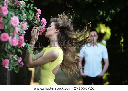 Sexual young blonde woman with bright makeup and curly hair in yellow dress smelling lush bush of pink rose and man in background, horizontal picture