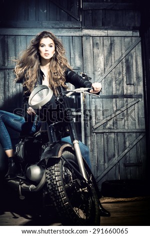 Cool sensual biker girl in black leather jacket and blue jeans sitting on old fashioned motorcycle in garage interior on grey wooden wall background, vertical picture
