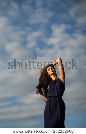 Young attractive sensual woman in long blue dress with curly hair standing on light blue sky and white clouds background copyspace, vertical picture