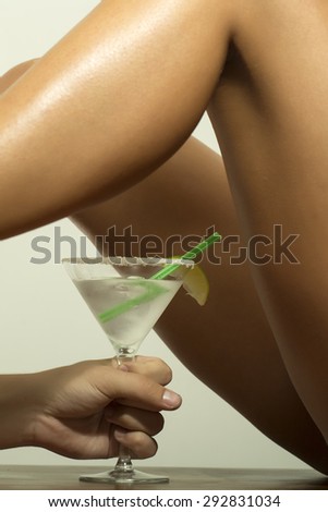 Slim naked sexy female legs and hand holding cocktail with lime in glass on white background, vertical picture