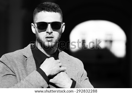 Handsome well dressed man in sunglasses straightens tie standing on outdoor background copyspace, black and white, horizontal picture