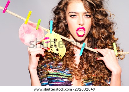 Portrait of sensual bright girl eating slice of cheese with pink lipstick kiss and bacon hanging on rope looking forward standing on grey wall background, horizontal picture