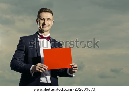Beautiful smiling man in formal suit holding orange sheet of paper standing on blue sky background copyspace, horizontal picture