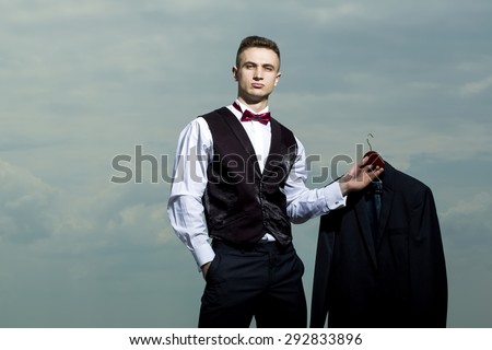 Handsome sexy brutal strong muscular man in formal suit with bow tie holding jacket standing on blue sky background copyspace, horizontal picture