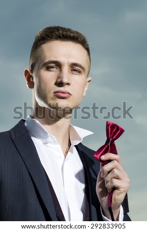 Portrait of handsome strong man in formal suit holding red bow tie standing on blue sky background, vertical picture