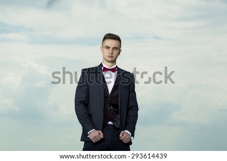 Attractive successful man in formal suit with bow tie standing on blue and white sky background, horizontal picture