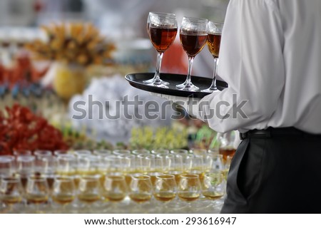 Back view of elegant waiter in formal black and white clothes with serving tray with wine glasses of amber hard beverage of cognac or brandy near full buffet table, horizontal picture