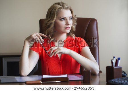 Beautiful sexy blonde business woman with curly hair in red blouse sitting in office at table with office supplies holding glasses, horizontal picture