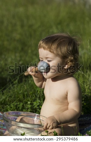 Small funny male child with curly hair sitting in lawn on green grass eating cream of wheat from glass plate with spoon sunny day, vertical picture