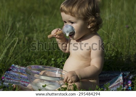 Small pretty male child with curly hair sitting in lawn on green grass eating cream of wheat from glass plate with spoon sunny day, horizontal picture