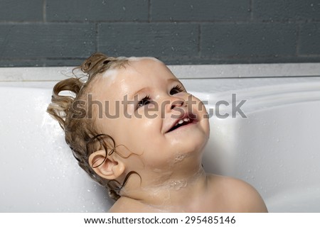 Small happy pretty baby boy sitting in white bathroom with wet foam hair looking away, horizontal picture