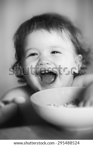 Portrait of funny little laughing baby boy with curly hair and round cheecks eating from plate black and white closeup, vertical picture