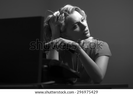 Overworked pretty secretary woman sitting at office table underlaying in blouse holding phone receiver on bare wall background black and white, horizontal picture