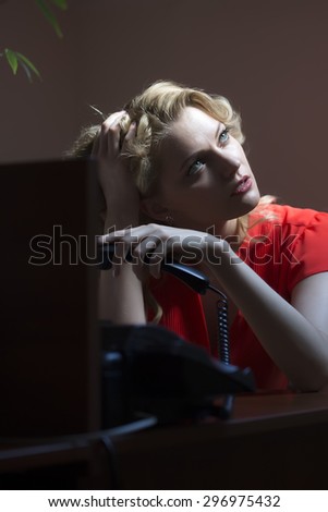 Pensive pretty secretary blond woman sitting at office table underlaying in red blouse holding phone receiver on bare wall background, vertical picture