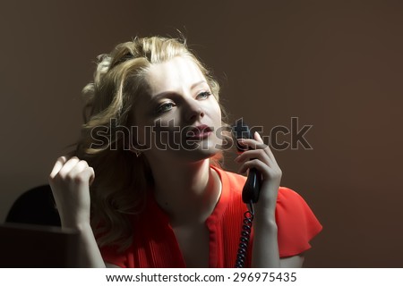 Working cute blonde secretary woman sitting at office table in red blouse holding telephone receiver calling someone on bare wall background, horizontal picture