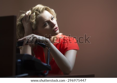 Bored attractive blonde secretary woman sitting at office table underlaying in red blouse holding phone receiver on bare wall background, horizontal picture