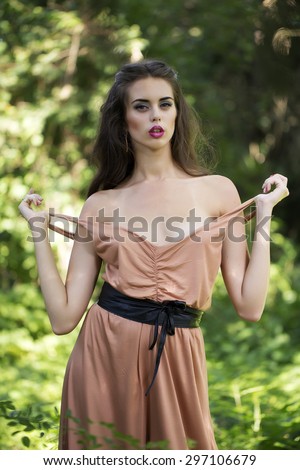 Attractive erotic sexy young woman with bright makeup and long curly brunette hair in beige dress and black belt standing and undressing in green garden on natural background, vertical picture