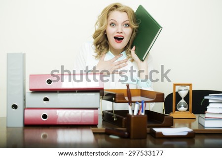 Pretty young secretary girl with curly hair has stress sitting at table with many folders documents and office appliances holding green notebook on white background, horizontal picture