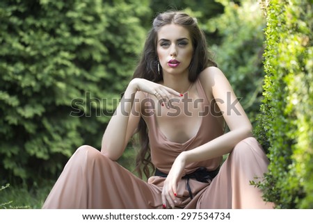 Pretty sensual young woman with bright makeup and long curly brunette hair in summer overalls with deep decollete sitting in green garden with fresh plants sunny day outdoor, horizontal picture