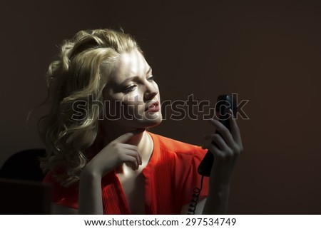 Serious beautiful blonde secretary woman sitting at office table in red blouse holding phone receiver calling someone on bare wall background, horizontal picture