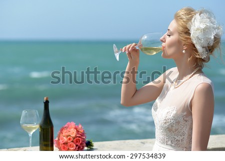 Beautiful sensual young bride in wedding dress standing on balcony drinking white wine from glass near bottle and rose bouquet sunny day on blue water and sky background, horizontal picture