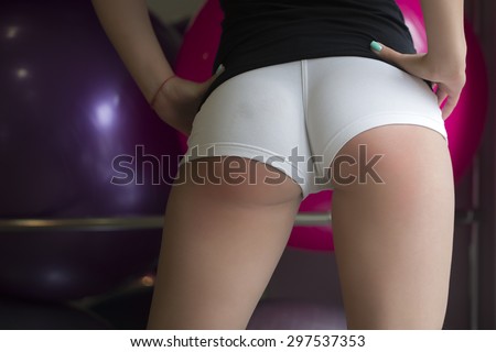 Closeup back view of beautiful sexy beefy female bottom with straight soft skinned legs in skin-tight white shorts standing in sport hall on big fitness balls background, horizontal picture
