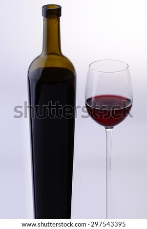Two objects oblong of green full bottle and glass goblet with red dessert wine standing together isolated on grey white studio background, vertical picture