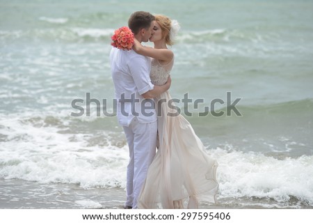 Beautiful young wedding couple of kissing man and woman in beige dress with rose bouquet emracing and standing on sea beach coast on wavy water background, horizontal picture
