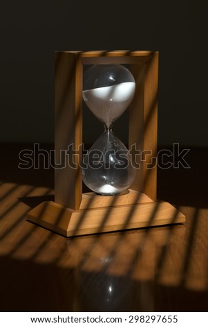 Old fashioned retro wooden hour glass clock with white sand standing on table top with jalousie shadow from sun through window, vertical picture