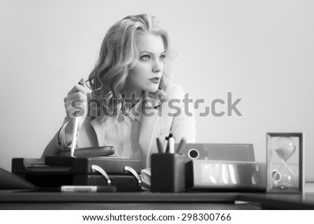 Beautiful pensive sexy business woman sitting at table with many office appliances holding knife for cutting paper looking away black and white, horizontal picture