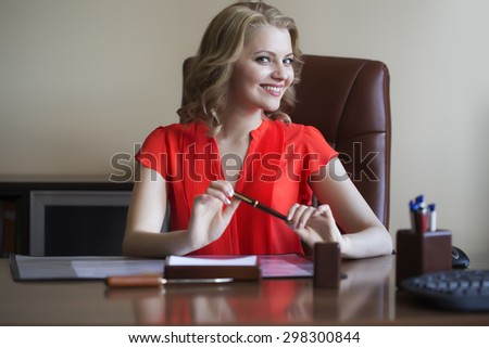Cute young elegant smiling business woman sitting in office on brown leather chair in red blouse holding pen in hands looking forward indoor on white background, horizontal picture