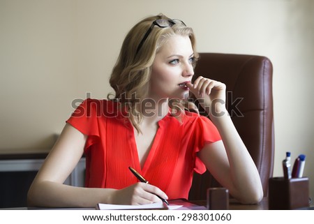 Attractive young elegant smart blonde working business woman sitting in office on brown leather chair in red blouse and glasses thinking and writing looking away indoor on white background