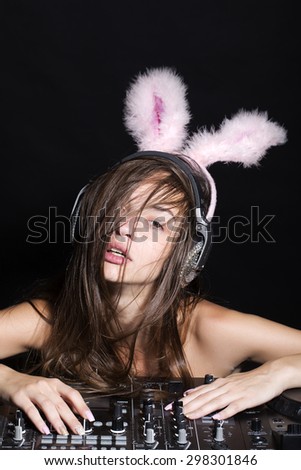 Portrait of young beautiful sexual dj woman in earphones and bunny ears with naked shoulders and long hair near professional musical mixer console on black studio background, vertical picture
