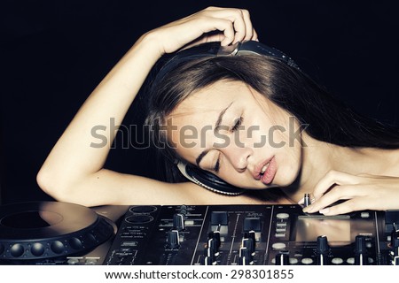 Portrait of young beautiful sexy disk jockey girl in headphones lying with closed eyes on professional musical mixer console on black studio background, horizontal picture