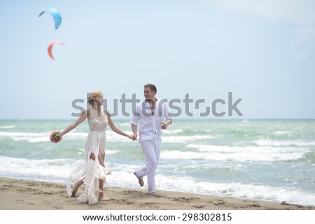 Temder beautiful young wedding couple of boy and girl in white running along ocean beach coast on windy weather sunny day with paraplanes on blue sky background, horizontal picture