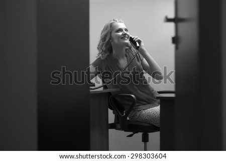 View from doorway of smiling beautiful secretary woman sitting in office with feet on table in blouse holding telephone receiver speaking on phone black and white, horizontal picture