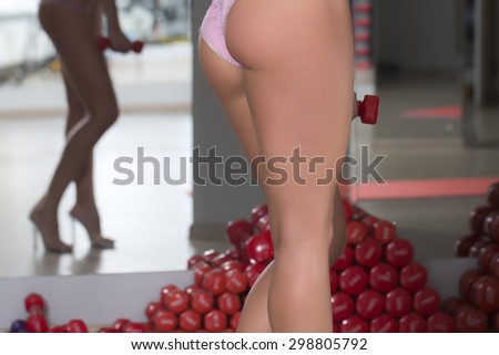 Closeup of beautiful sexual female bum with straight legs and hips in pink glamour panties standing in sport fitness hall near heap of red dumbells on mirror reflection background, horizontal picture