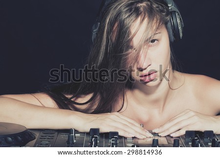 Portrait of young beautiful sexual disk jockey woman in headphones with naked shoulders near professional musical mixer console on black studio background, horizontal picture