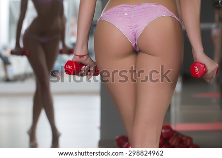 Closeup of beautiful sexy beefy female bum with straight legs in pink glamour bikini standing in sport fitness hall holding red dumbells on mirror reflection background, horizontal picture