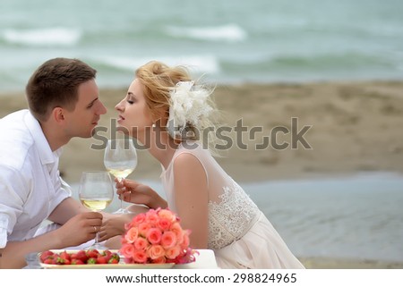 Beautiful tender wedding couple of young man and woman sitting on ocean beach shore at table with rose bouquet red strawberry kissing and drinking white wine from glasses copyspace, horizontal picture