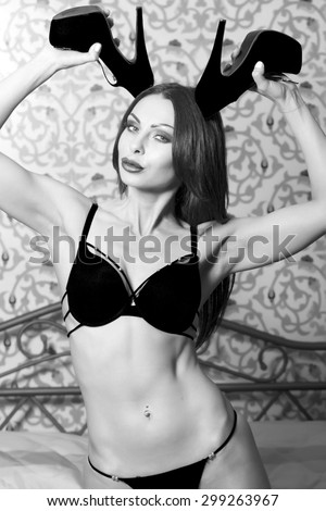 Sexual pretty young woman in erotic underwear sitting on bed in bedroom holding chamois shoes on high heels as antlers looking forward on patterned wall background black and white, vertical picture