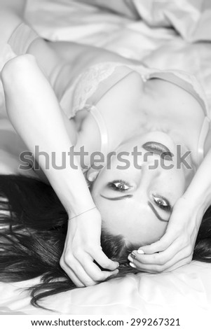 Beautiful sexy brunette woman with straight body and chest in erotic lace underwear lying on bed holding head with hands looking forward black and white, vertical picture