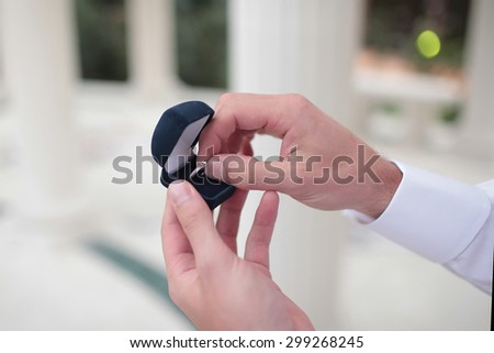 Man in white shirt takes engagement platinum ring with gemstone from open square dark blue box outside, horizontal photo