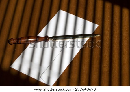 Elegant sharp golden paper knife with brown wooden handle lying on one white envelope on office table on jalousie shadow background, horizontal picture