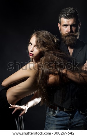 Young pair of sexual pretty girl with straight body with naked back and long hair standing near unshaven man with beard and moustache in waistcoat on black background copyspace, vertical picture