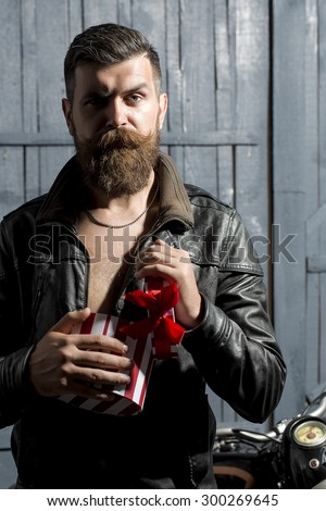 Handsome male biker with beard and moustache in brown leather jacket and chain holding open round red white striped present box with ribbon bow standing on grey wooden background, vertical picture