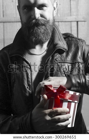 Attractive unshaven guy with beard and moustache in leather jacket and chain opening round red white striped gift box with ribbon bow standing on wooden background black and white, vertical picture