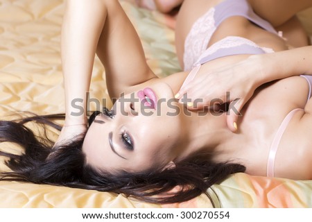 Pretty sexy brunette woman with straight body in erotic lace purple and lilac underwear lying on bed holding hands near face looking away, horizontal picture