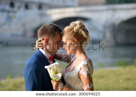 Tender young wedding pair of man in blue jacket and red tie bow embracing girl in white dress with posy of calla flowers standing near river and bridge on sunny outdoor background, horizontal picture