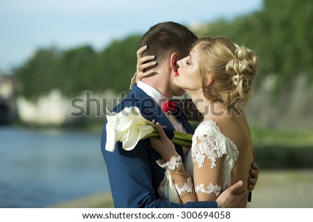 Sweet young wedding pair of man in blue jacket and red tie bow embracing blonde woman in white dress holding bouquet of calla flowers outdoor standing on sunny natural background, horizontal picture